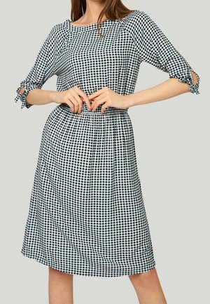 Donna che indossa un vestito a quadri bianco e nero lungo fino al ginocchio con maniche a tre quarti annodate ai gomiti su uno sfondo semplice.