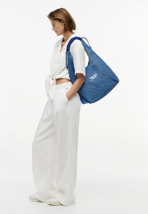 Donna con abito bianco che porta una grande borsa Tommy Jeans blu testurizzata sulla spalla, in piedi di profilo su sfondo bianco.