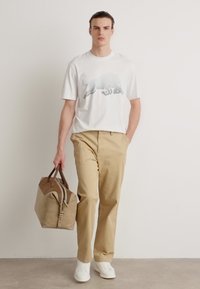 Hvit t-skjorte med grafikk av en bjørn i gråtoner, kombinert med khaki bukser og en tan duffelbag med mørke detaljer. Hvite sneakers.