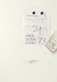 Tarjeta de medidas hecha de papel blanco, con texto en negro que muestra números para el tamaño de hombros, longitud total, longitudes de las mangas interior y exterior, adjunta a una pared.