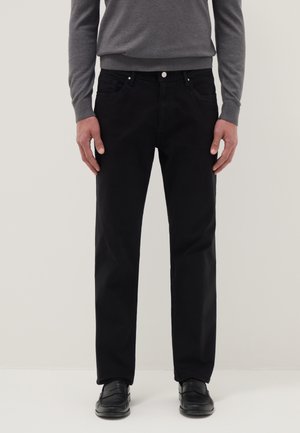 Uomo con pantaloni neri a gamba dritta, maglione grigio scuro a maniche lunghe e mocassini neri, in piedi contro uno sfondo semplice.