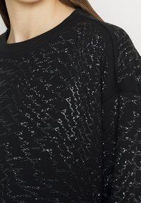 Sudadera negra con detalles plateados brillantes en un patrón ondulado. Tejido suave con un cuello redondo y corte relajado.
