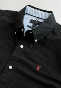 Schwarzes Hemd mit Knopfleiste in schlanker Passform. Verfügt über eine Krageninnenseite in Blau und Weiß gestreift, weiße Knöpfe und ein kleines rotes gesticktes Emblem.