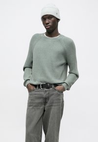 Gaiši zaļa ribaina džemperis ar apaļu kakla izgriezumu un garām piedurknēm, kopā ar pelēkām džinsa biksēm un gaiši pelēku adītu cepuri.