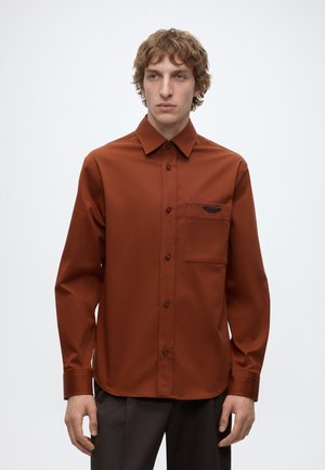 Homme portant une chemise à manches longues couleur rouille avec une poche poitrine et un pantalon sombre, debout devant un fond clair uni.