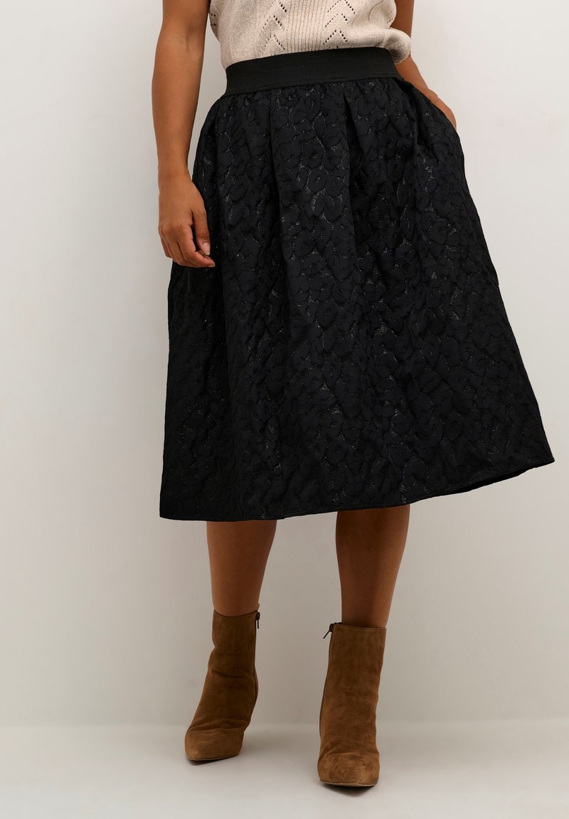 Femme portant une jupe midi noire texturée avec des poches, un haut en maille beige et des bottines en daim marron, debout devant un fond uni.