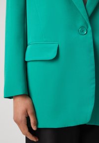 Blazer en verde azulado con cierre de un solo botón, tejido suave, solapa con muesca y un bolsillo de tapa. El brazo es parcialmente visible, con tela negra debajo.
