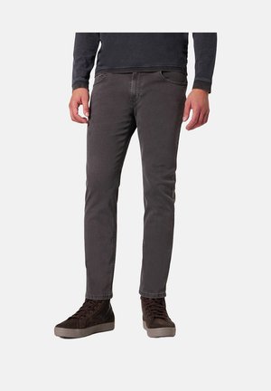Mann trägt dunkelgraue Slim-Fit-Jeans und dunkelbraune High-Top-Sneakers und steht vor einem weißen Hintergrund.