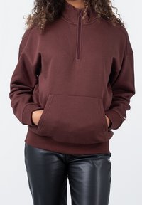 Brązowy sweter z półzamkiem, z długimi rękawami, bocznymi kieszeniami i luźnym krojem. Materiał wydaje się być miękki i lekko teksturowany.