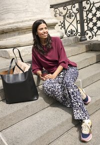 Donna sorridente in pantaloni con fantasia e maglione color bordeaux seduta su gradini di pietra, tiene una tazza di caffè con una borsa nera e un giornale accanto a lei.