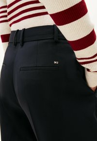 Pantaloni neri su misura con una superficie liscia, dotati di una tasca posteriore e un piccolo logo decorativo. Abbinati a un maglione a righe con lavorazione a costine.