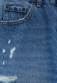 Gros plan sur un tissu en denim bleu montrant une poche avant avec des coutures orange et de petites déchirures usées près de la poche et dans la partie inférieure.