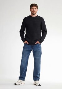 Zwarte gebreide trui met textuur, blauwe spijkerbroek en witte sneakers. De trui heeft lange mouwen en een ronde hals. De spijkerbroek is loszittend.