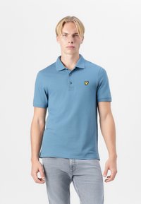 Lyseblå polo skjorte med to knapper og korte ermer; har en liten gul logo på venstre bryst. Glatt bomullsstoff.