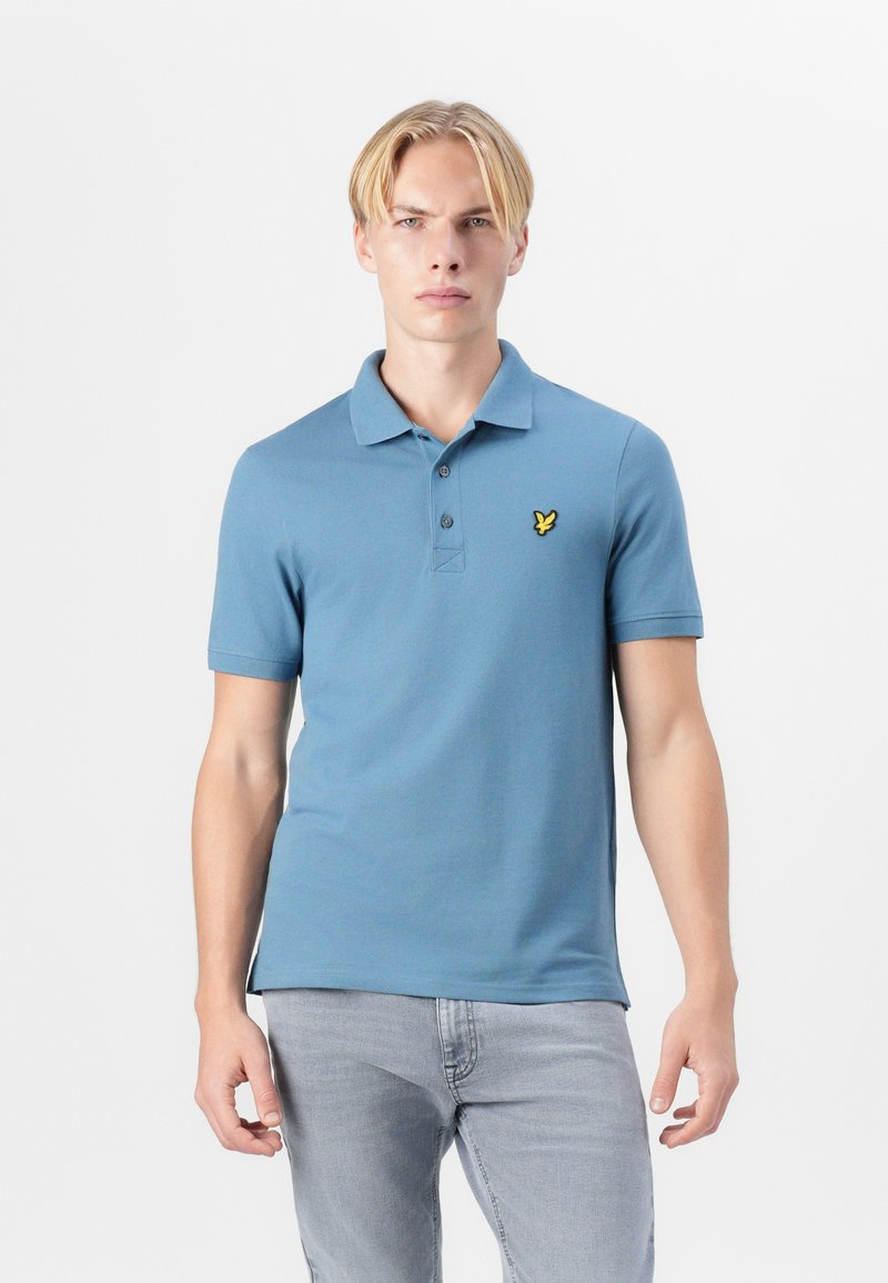 Lyseblå polo skjorte med to knapper og korte ermer; har en liten gul logo på venstre bryst. Glatt bomullsstoff.