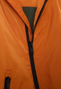 Chaqueta ligera naranja con un zipper negro, que cuenta con un forro de polar verde y un bolsillo lateral con un zipper negro oculto.