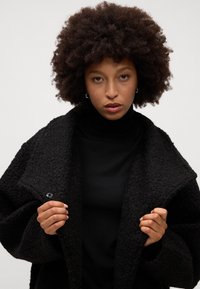 Cappotto oversize nera a trama con colletto sciallato, caratterizzato da una chiusura con un singolo bottone, indossato sopra un dolcevita nera.