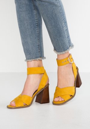 Pieds vêtus de sandales à talon bloc en daim jaune avec brides autour de la cheville et boucles, associées à un jean bleu court à ourlet effiloché.