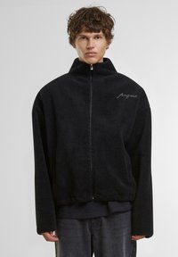 Veste zippée en polaire noire avec col haut, coupe oversize et subtil détail de logo brodé sur la poitrine gauche. Tissu texturé.