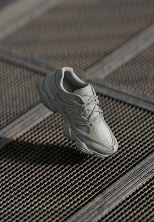 Baskets athlétiques gris clair avec des détails en mesh et synthétiques reposant sur une surface en grille métallique texturée.