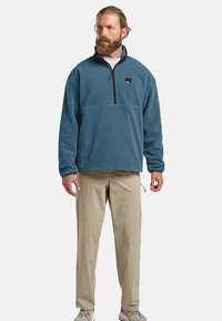 Fleece pullover in tealblauw met een ritssluiting bij de hals, een losse pasvorm en een klein logoplaatje op de borst, gecombineerd met beige broek.