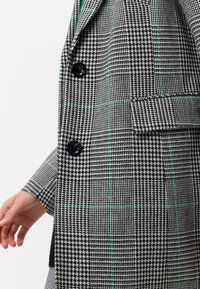 Cappotto doppiopetto in cashemere a scacchi bianco e nero con dettagli a quadretti verdi. Presenta bottoni neri e tessuto testurizzato.