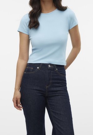 Vrouw in een lichtblauwe korte mouwen top en donkere high-waisted jeans voor een effen achtergrond.
