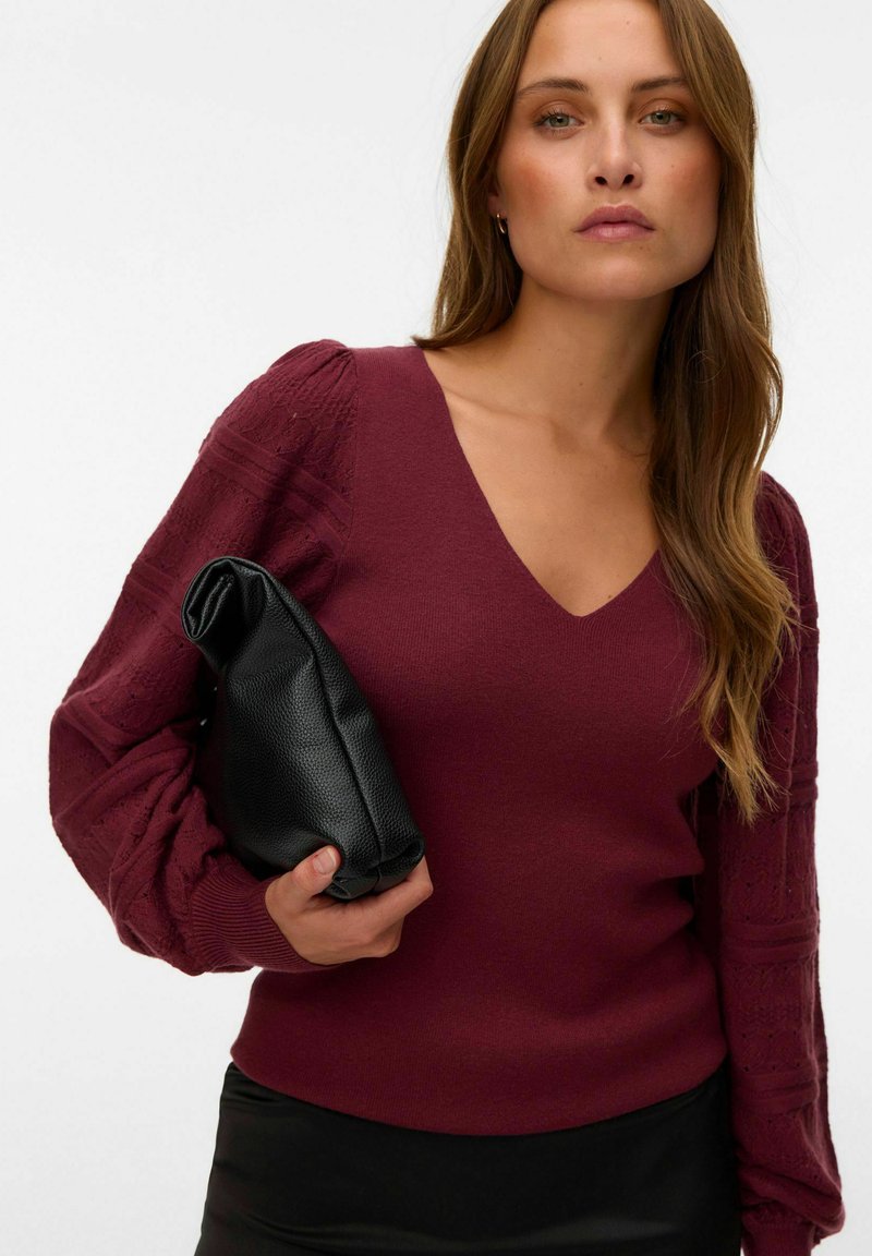 Femme portant un pull bordeaux à col V avec des manches texturées tient une pochette en cuir noire sur un fond blanc uni.