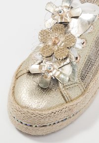 Zapato texturizado en dorado con adornos florales y acentos metálicos. Presenta una suela de yute trenzado con detalles iridiscentes.