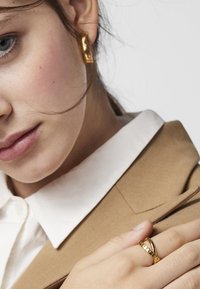 Złote kolczyki o prostokątnym kształcie, noszone razem ze złotym pierścionkiem o teksturowanej powierzchni. Widoczna jest jasnobrązowa marynarka z białym kołnierzykiem.