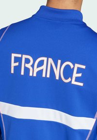Veste de sport bleue arborant le texte "FRANCE" en lettres orange surlignées et une bande horizontale blanche sur le dos.
