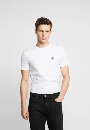 Jeune homme aux cheveux bouclés portant un t-shirt blanc à manches courtes avec un petit logo "cK" et un jean noir, debout devant un fond gris uni.