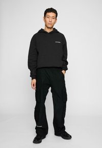 Czarna bluza z kapturem z przednią kieszenią i białym napisem, w połączeniu z czarnymi spodniami cargo z bocznymi kieszeniami, obie w luźnym kroju. Czarne sneakersy.