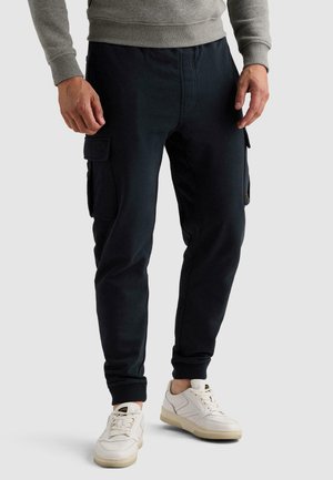 Άνδρας που φοράει μαύρο cargo παντελόνι τύπου jogger και λευκά αθλητικά παπούτσια στέκεται με τα χέρια χαλαρά δίπλα στο σώμα σε απλό φόντο.