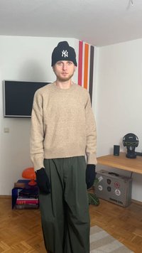 Béžový vlněný svetr, volný střih, žebrovaný lem; tmavě zelené široké kalhoty; černé rukavice; černá čepice s logem; minimalistické vnitřní pozadí.