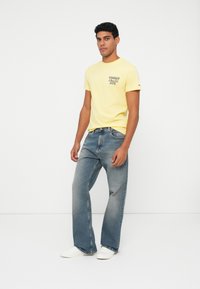 Mees seisab, seljas kollane Tommy Jeans t-särk, sinised teksad ja valged tossud, taustal lihtne valge taust.