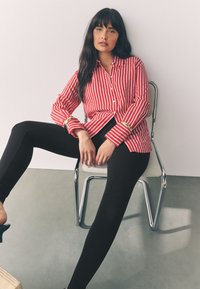 Rød- og hvidstribet skjorte med knapper, lange ærmer og gyldne detaljer, kombineret med sorte leggings. Person siddende på en metalstol.