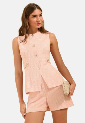 Naine, kes kannab roosakas-halli varrukateta tekstuuriga nööpidega topi ja sobivaid pükse, hoides väikest beeži kootud käekotti.