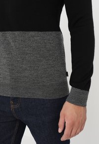 Strickpullover mit einem schwarzen oberen Abschnitt und einer grauen unteren Hälfte. Verfügt über gerippte Bündchen und eine glatte Textur. Sichtbares angebrachtes Logoetikett.