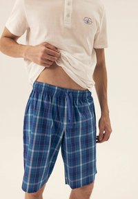Blaue karierten Shorts mit einem Zugbandbund, die verschiedene Blautöne und weiße Linien aufweisen. Darüber wird ein eng anliegendes weißes Polohemd getragen.