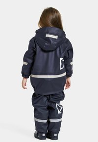 Marinblå vattentät jacka och byxor med reflexremsor, huva och elastiska muddar; har en logotyp på ryggen och en slät textur.