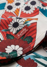 Tissu floral en rouge, blanc et turquoise avec une surface texturée. Comprend une étiquette rouge avec "Superdry Édition Spéciale" et divers motifs de fleurs.