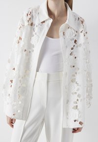 White floral lace shirt with a collared neckline, featuring textured detailing and a button-down front, paired with white trousers.