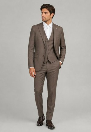 Homme portant un costume trois pièces marron avec une chemise blanche et des chaussures habillées marron foncé, debout avec une main dans sa poche.