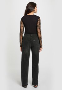 Kvinde der står med ryggen til, iført en sort top med gennemskinnelige blonde lange ærmer og højtaljede løstsiddende sorte jeans med baglommer.