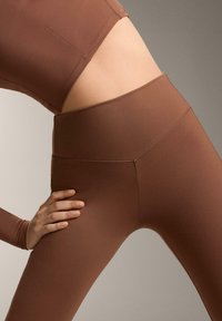 Brązowe, elastyczne legginsy wykonane z gładkiej, lekkiej tkaniny. Posiadają wysoki pas i bezszwową konstrukcję z minimalną ilością szwów.