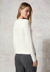 Weiße Langarmbluse mit Rundhalsausschnitt und dezenten Nahtdetails, kombiniert mit dunkelgrauen Hosen. Glatter Stoff, lockerer Schnitt.