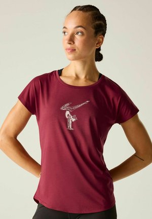 Woman with braided hair wearing a burgundy t-shirt with a yoga pose graphic and text "Stretch Yoga" stands with hands on hips looking sideways.