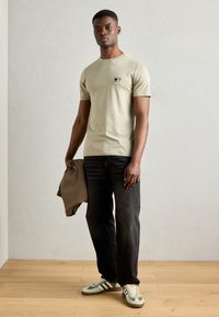 Gai olīvu zaļas krāsas T-krekls ar nelielu dzēriena grafiku, kombinēts ar tumšiem džinsiem un zaļi svītrainiem sporta apaviem; ikdienas stils, dabiska auduma tekstūra.