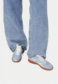 Asmuo, vilkintis šviesiai mėlynus "Reebok" sportbačius su baltais raišteliais ir guminiu padu, derinamas su laisvai krentančiomis šviesiai mėlynomis džinsinėmis kelnėmis.